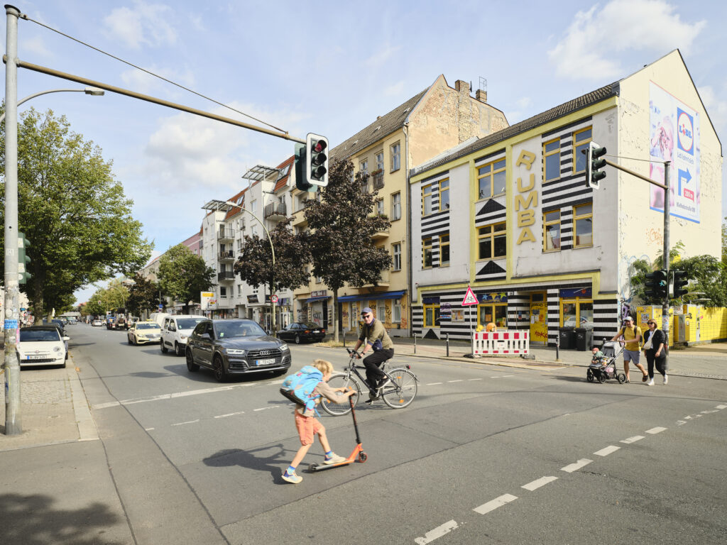Auf dem Bild erkennbar ist eine Straßenkreuzung, auf der Personen über eine Ampel laufen. Zu sehen ist eine Häuserzeile, auf dem ersten Haus steht der Name Rumba. Autos halten an der Ampel.