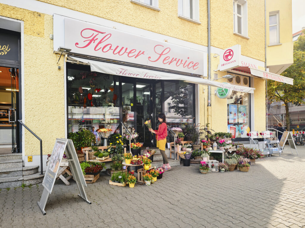 Auf dem Bild zu sehen sind Erdgeschossläden in einem gelben Haus. Links sieht man einen Laden mit dem Namen Flower Service, vor dem Pflanzen stehen und rechts an der Ecke des Hauses befindet sich Rossmann.