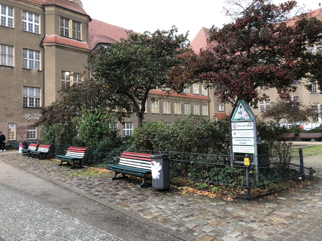 Das Bild zeigt den begrünten Vorplatz der Volkshochschule Treptow-Köpenick in der Baumschulenstraße. Zu sehen sind Bäume, Bänke sowie das Gebäude der Volkshochschule im Hintergrund.