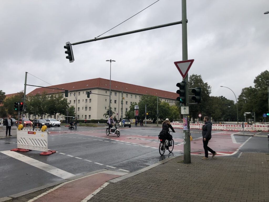 Das Bild zeigt den Straßenraum der Köpenicker Landstraße an einer Kreuzung mit einer Ampel, Radfahrenden und Fußgängern.