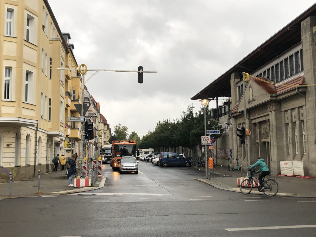 Das Bild zeigt den Straßenraum an der Ecke Glanzstraße/Baumschulenstraße mit parkenden Autos und einem Fahrradfahrer.